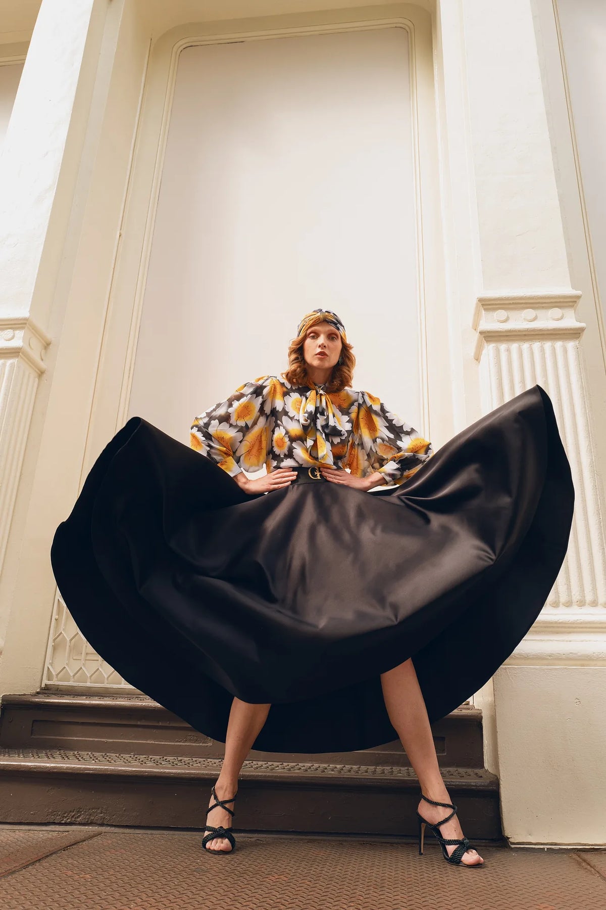 FRAQAIR daisy print blouse and turban with a black satin circle skirt held wide, on the steps of a cream colonnaded building