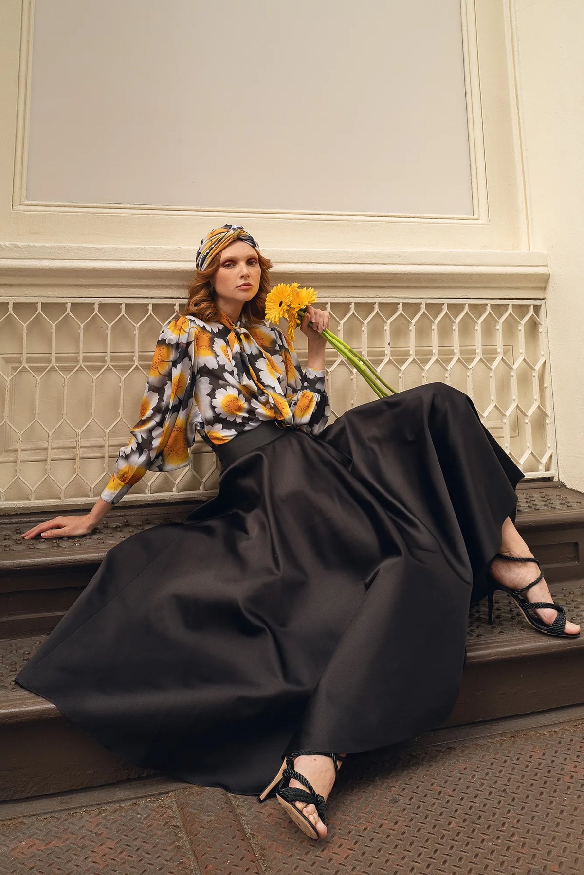 FRAQAIR daisy print blouse and turban with a black satin circle skirt, seated on the steps with yellow gerberas